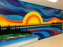 photo shows a mural painted in the hallway of Gypsumville School, MB. It shows a lake, tree line and sky, the skyline depicts waves of colour showing the 7 sacred teachings symbols