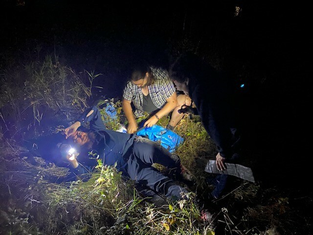 photo depicts a man laying on the ground holding a flashlight while a woman assists him in first aid. The photo is taken in the dark.